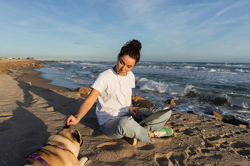 young woman in wired earphones sitting with laptop and cuddling pug dog on beach in Barcelona