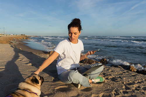 young woman in wired earphones holding smartphone near laptop and cuddling pug dog on beach in Barcelona