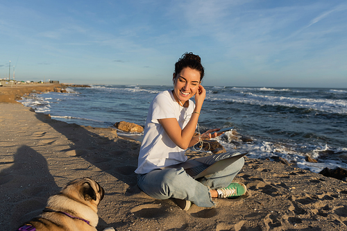 happy woman wearing wired earphones while holding smartphone near laptop and pug dog on beach in Barcelona