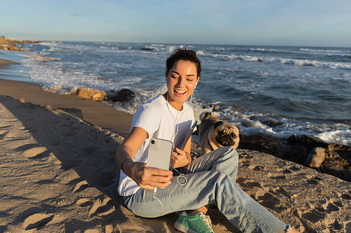 happy woman in wired earphones holding laptop and taking selfie with pug dog on beach in Barcelona
