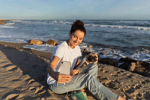cheerful woman in wired earphones holding laptop and taking selfie with pug dog near sea in Spain
