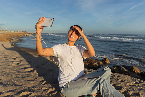 cheerful woman in wired earphones sitting with laptop and taking selfie near sea in Spain