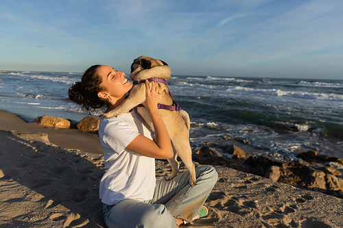 positive young woman with closed eyes holding pug dog on beach near sea in Barcelona