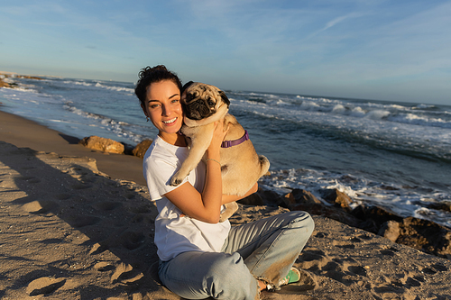 positive young woman with curly hair holding pug dog on beach near sea in Barcelona