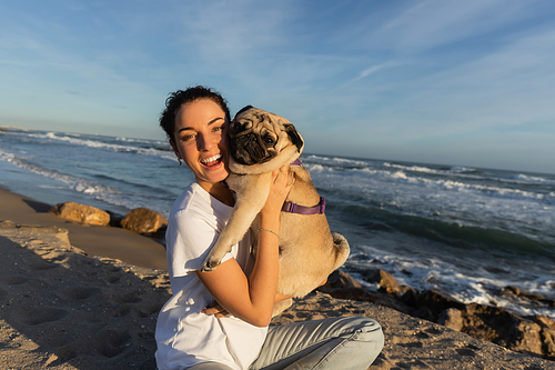 joyful young woman with curly hair holding pug dog on beach near sea in Barcelona