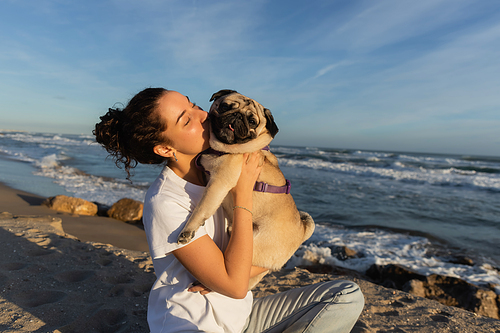 young woman with curly hair kissing pug dog on beach near sea in Barcelona