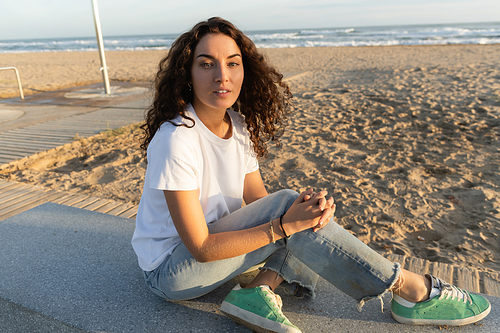 pretty woman with curly hair sitting on sandy beach near sea in Barcelona
