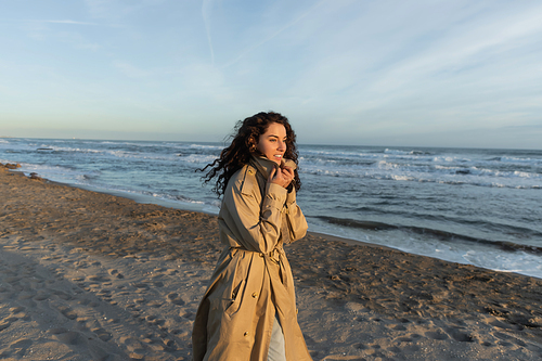 cheerful woman in stylish beige trench coat walking on beach in Barcelona