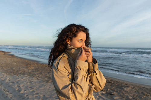curly young woman covering face with collar of beige trench coat on beach in Barcelona