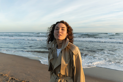 curly young woman in beige trench coat and wired earphones listening music near sea in Barcelona