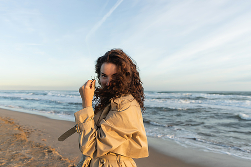 young woman in beige trench coat adjusting curly hair while looking at camera near sea in Barcelona