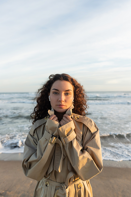 curly young woman in wired earphones listening music while adjusting collar of beige trench coat near sea in Barcelona