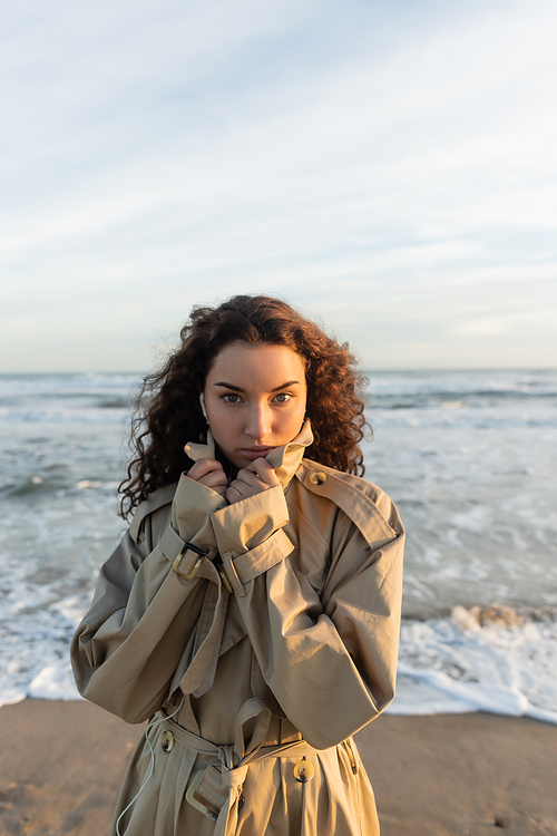 curly woman in wired earphones listening music while adjusting collar of beige trench coat near sea in Barcelona
