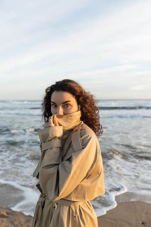 curly woman in wired earphones adjusting collar of beige trench coat and looking at camera near sea in Barcelona