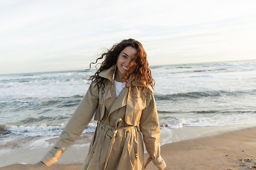 cheerful young woman in beige trench coat and wired earphones listening music near sea in Barcelona
