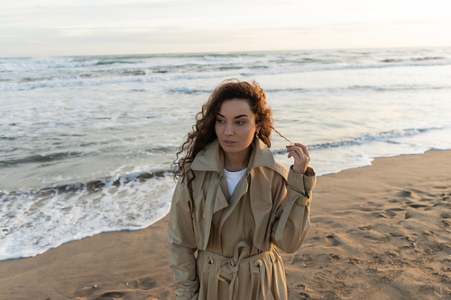Young woman in trench coat touching curly hair on beach