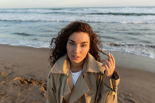 Curly woman in trench coat looking at camera on blurred beach near sea
