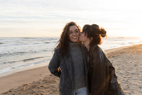 Young woman kissing cheek of smiling friend on beach in Barcelona