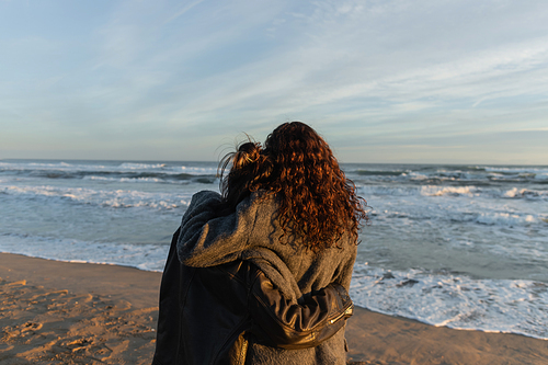 Back view of friends hugging on beach near sea in Spain