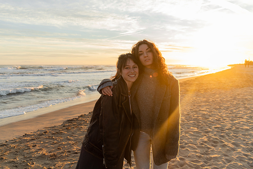 Curly woman hugging smiling friend on beach during sunset in Barcelona