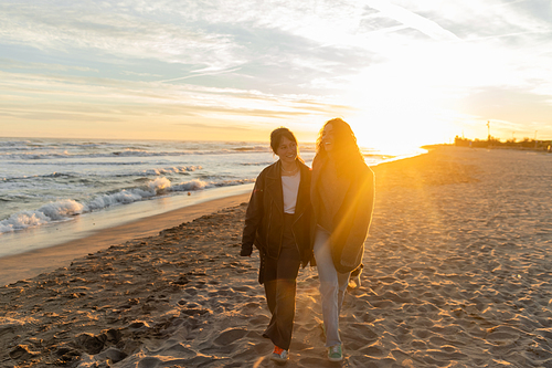 Positive women walking on beach near sea during sunset