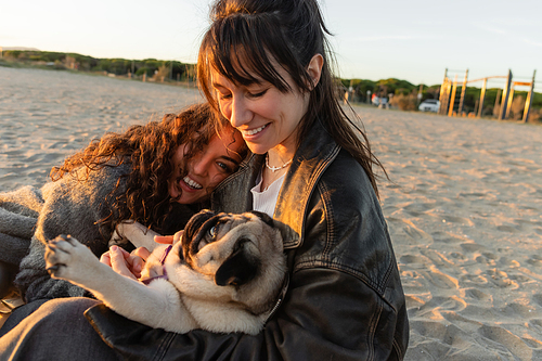 Positive young women playing with pug dog on beach in evening