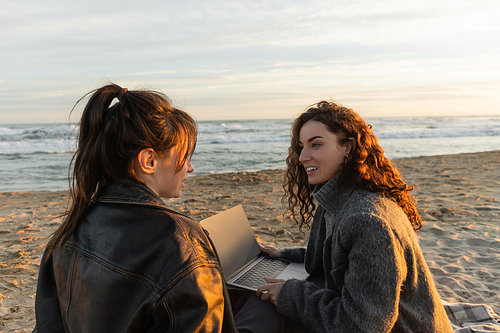 Smiling woman holding laptop while talking to friend on beach in evening