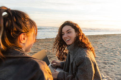 Smiling curly woman using laptop and talking o friend on beach