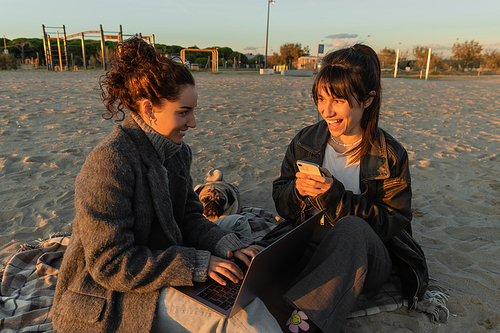 Cheerful women using gadgets near pug dog on beach in Spain