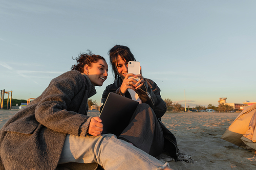 Smiling friends using smartphone and laptop on beach in Spain