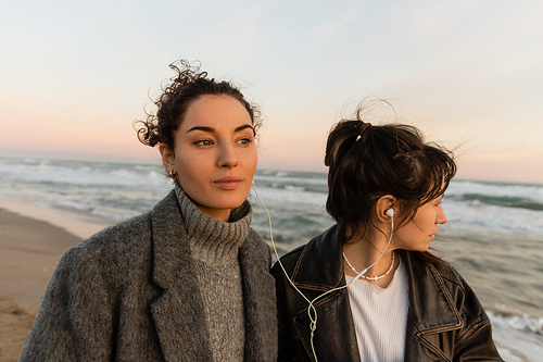 Curly woman listening music in wired earphones with friend on beach in Barcelona