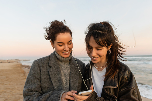 Positive friends in wired earphones using cellphone on beach in Spain