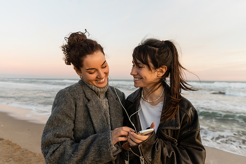 Smiling friends listening music in wired earphones and holding smartphone on beach