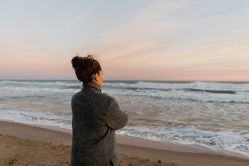 Curly woman in coat looking at sea while standing on beach in Barcelona