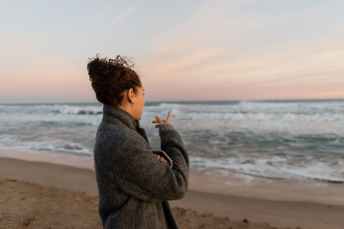 Curly woman in coat pointing at sea while standing on beach