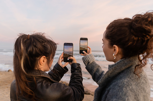 Young friends taking photo on smartphones while standing on beach in Spain