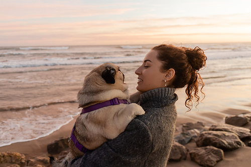 Side view of smiling curly woman in coat holding pug dog on beach in Barcelona