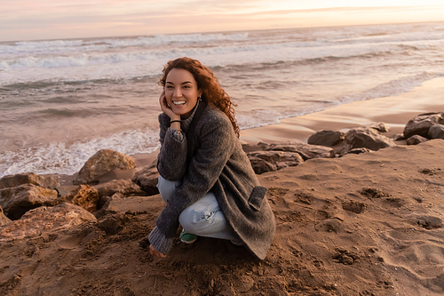 Positive woman in coat looking at camera near stones on sandy beach near sea