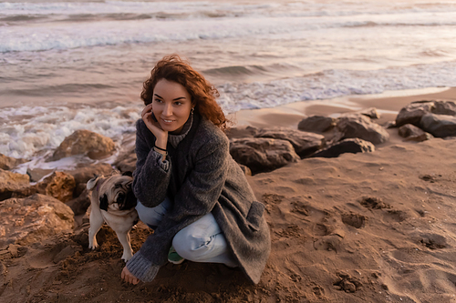 Smiling woman in coat looking away near pug dog on beach in Barcelona