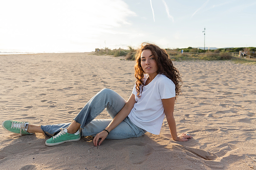 Curly woman in jeans and t-shirt sitting on beach in Spain