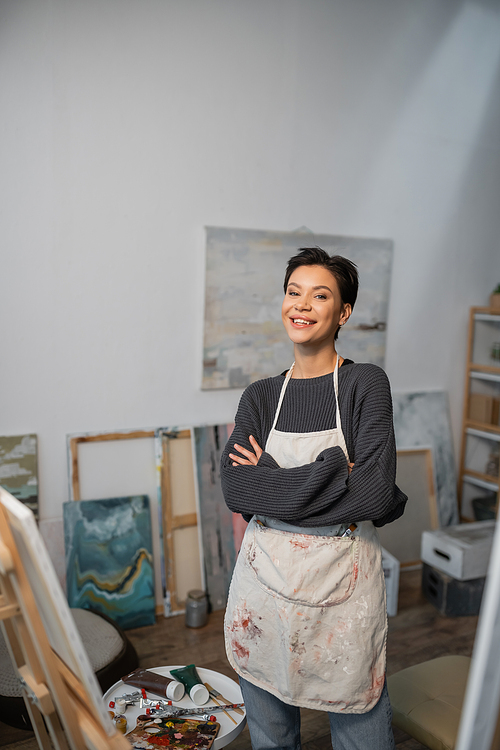 Cheerful artist in apron looking at camera near paints and easel in studio