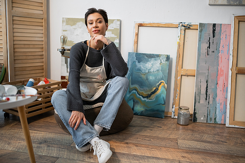 Tattooed artist in apron sitting on floor near paintings in studio