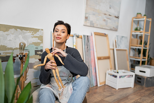 Young artist holding wooden doll and looking at camera near paintings in studio