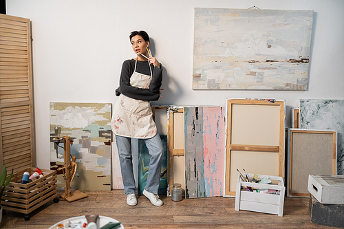 Dreamy artist holding paintbrushes while standing near drawings in studio