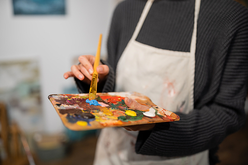 Cropped view of blurred artist in apron holding palette and paintbrush in studio