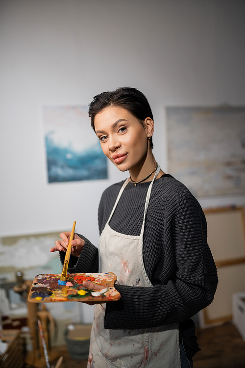 Portrait of short haired artist in apron holding paintbrush and palette in workshop