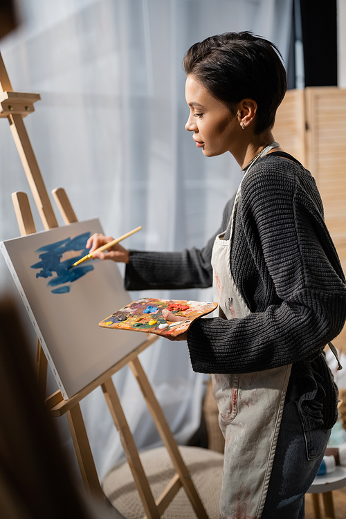 Side view of brunette artist painting on canvas in studio