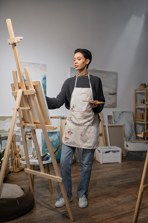Artist in apron holding palette and looking at canvas on easel in studio