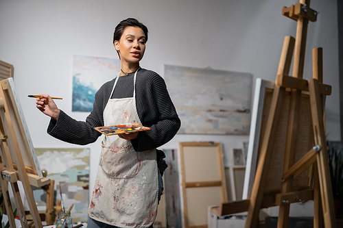 Short haired artist in apron holding paintbrush and palette near canvases on easels in studio