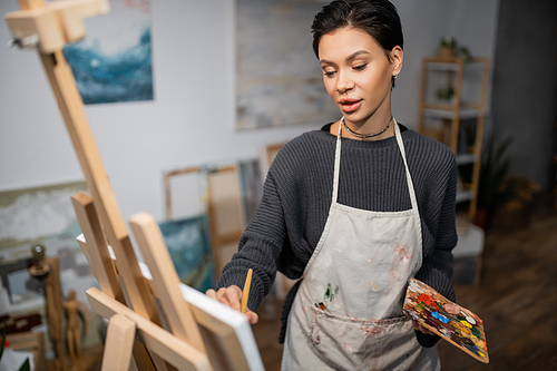 Short haired artist in apron holding palette while painting on blurred canvas